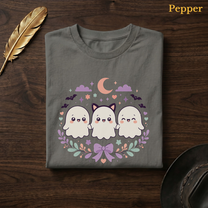 Spooky cute tshirt in Pepper, a warm grey-charcoal garment-dyed shirt folded flat on dark walnut wood beside a gold feather and a leather hat, the Ghoul Friends design centred on the chest, labelled Pepper.