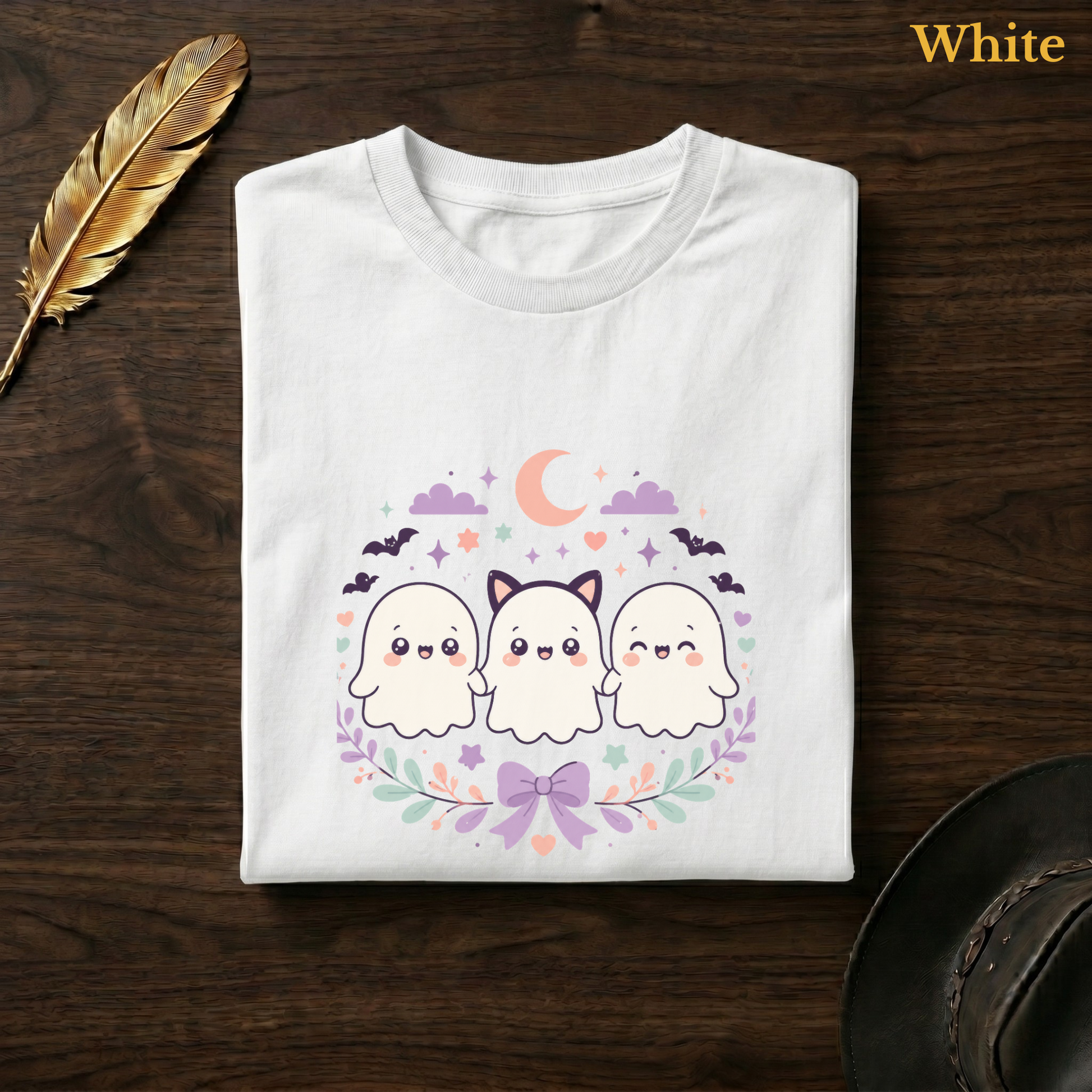 Ghost trio shirt in White, folded flat on dark walnut wood beside a gold feather and a leather hat, the full-colour kawaii ghost trio and cat-ear design printed on the chest, labelled White.