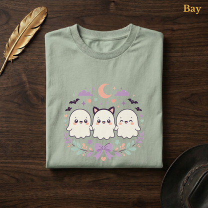 Ghoul Friends tee in Bay, a soft sage-green garment-dyed shirt folded flat on dark walnut wood beside a gold feather and a leather hat, the kawaii ghost trio design printed on the chest, labelled Bay.