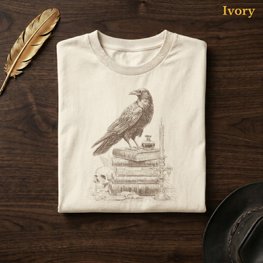 Poetcore shirt in Ivory, folded flat on dark walnut wood beside a gold feather and a leather hat, the engraved raven, inkwell, and skull design on the chest, labelled Ivory.
