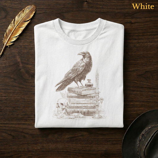 Raven scholar tee in White, folded flat on dark walnut wood beside a gold feather and a leather hat, the full woodcut-style raven, candelabra, and book stack design printed on the chest, labelled White.
