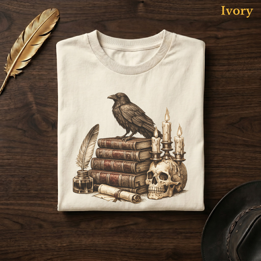 Poetcore apparel tee in Ivory, folded flat on dark walnut wood beside a gold feather and a leather hat, the raven, leather books, quill, and skull candelabra design on the chest, labelled Ivory.
