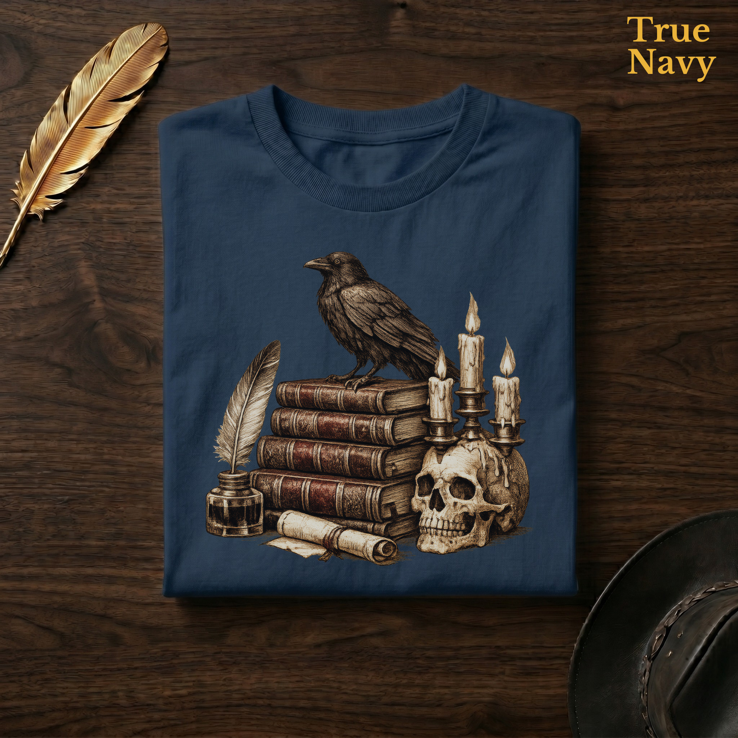 Poe's Study tee in True Navy, a deep navy garment-dyed shirt folded flat on dark walnut wood beside a gold feather and a leather hat, the raven and skull candelabra design on the chest, labelled True Navy.
