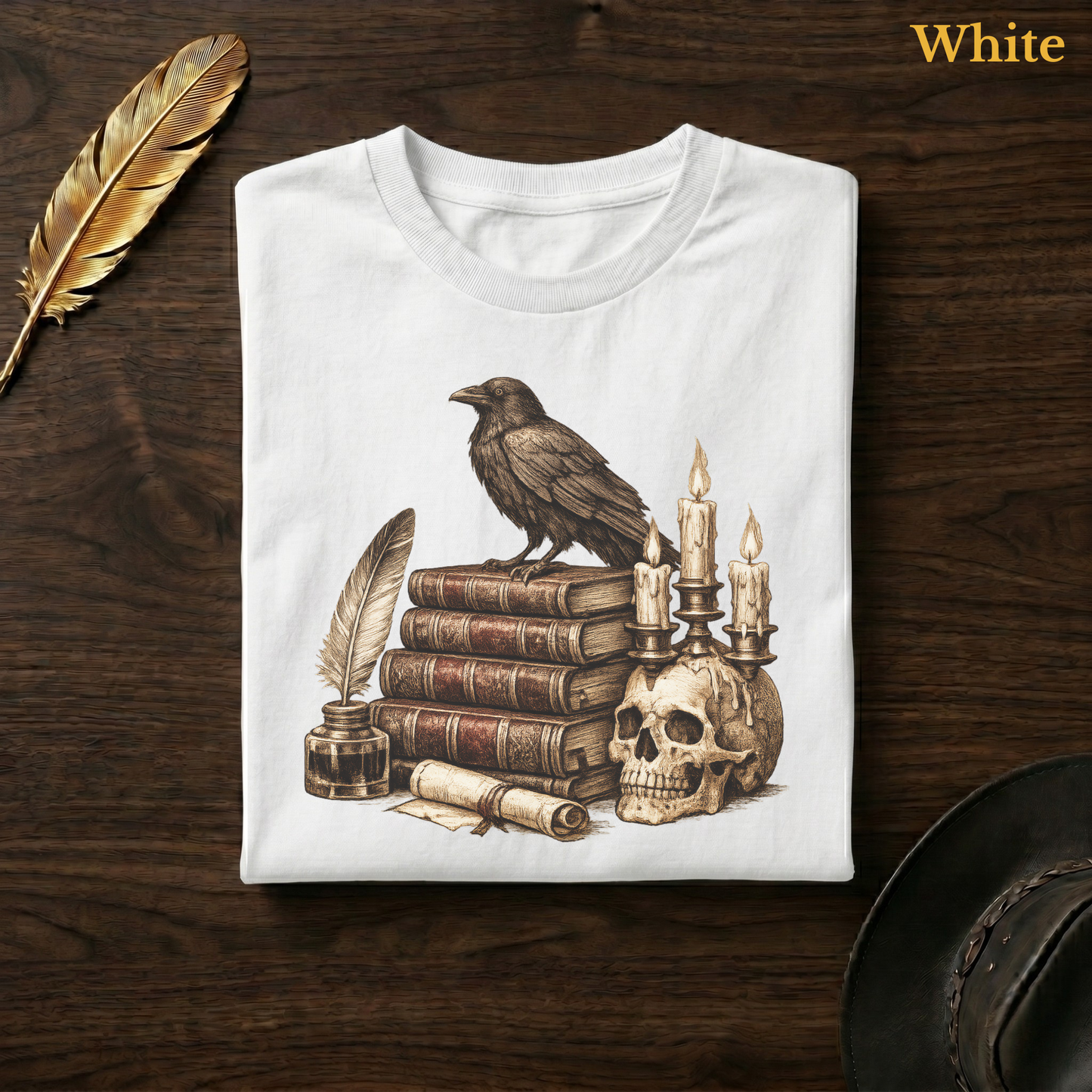 Nevermore raven tee in White, folded flat on dark walnut wood beside a gold feather and a leather hat, the full-detail sepia raven, books, quill, and skull candelabra design printed on the chest, labelled White.
