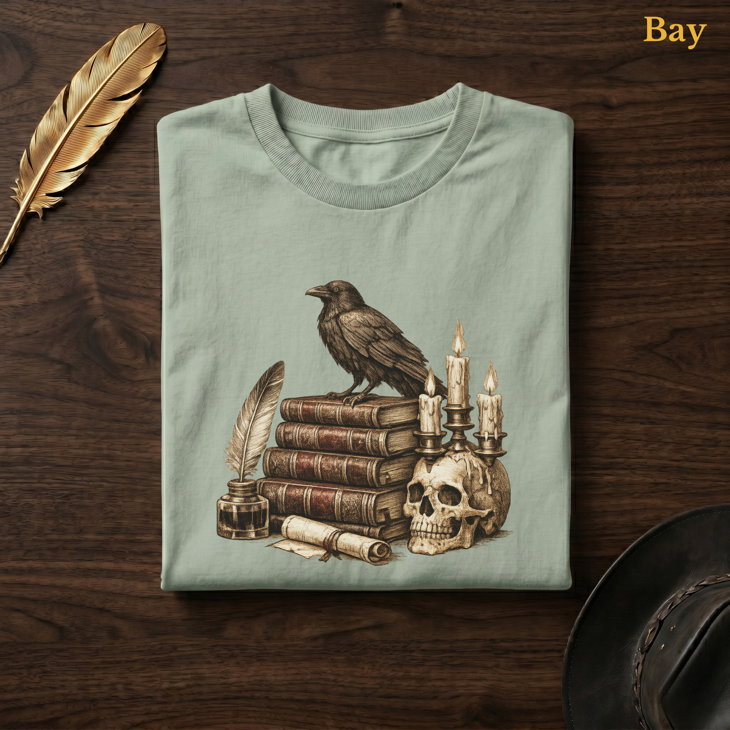 Poe's Study tee in Bay, a soft sage-green garment-dyed shirt folded flat on dark walnut wood beside a gold feather and a leather hat, the raven and skull candelabra design printed on the chest, labelled Bay.
