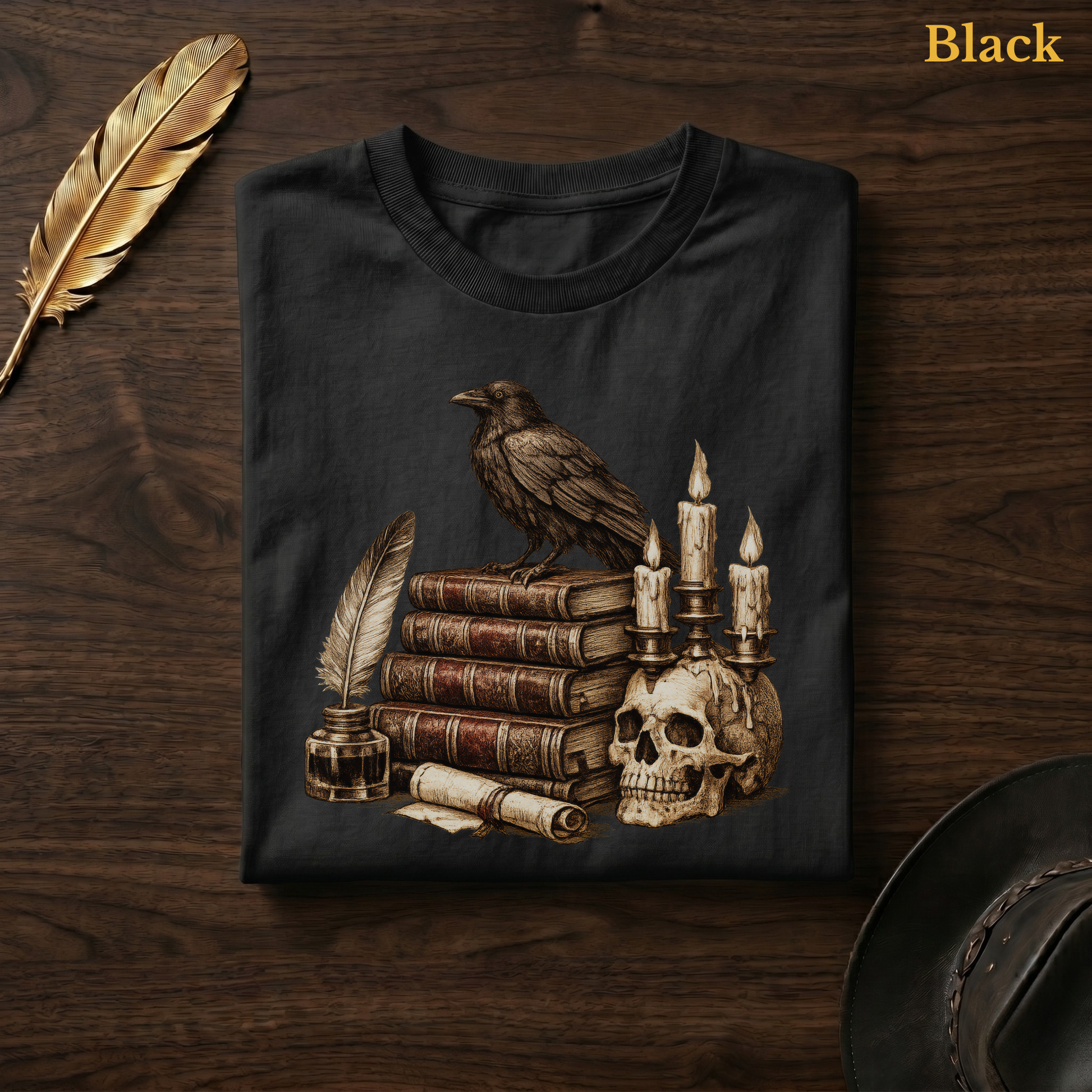 Poe's Study tee in Black, folded flat on dark walnut wood beside a gold feather and a leather hat, the engraved raven, books, quill, and skull candelabra design printed on the chest, labelled Black.
