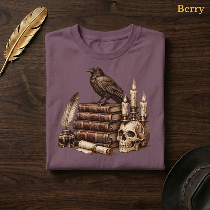 Poe's Study tee in Berry, a muted mauve-plum garment-dyed shirt folded flat on dark walnut wood beside a gold feather and a leather hat, the raven, books, and candelabra design on the chest, labelled Berry.
