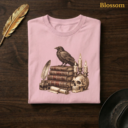 Poe's Study tee in Blossom, a soft pastel pink garment-dyed shirt folded flat on dark walnut wood beside a gold feather and a leather hat, the dark academia raven design on the chest, labelled Blossom.

