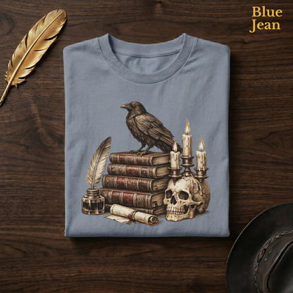 Bookish gothic tee in Blue Jean, a faded denim-blue garment-dyed shirt folded flat on dark walnut wood beside a gold feather and a leather hat, the Poe's Study raven and candelabra design on the chest, labelled Blue Jean.
