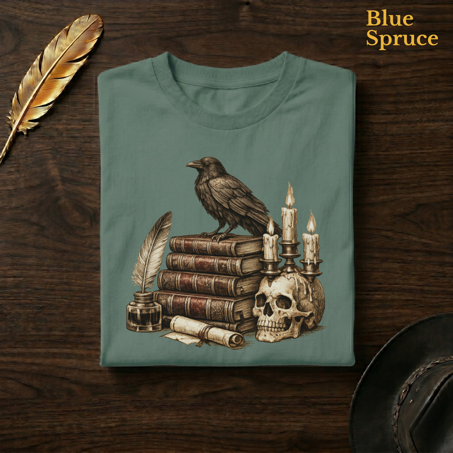 Gothic library tee in Blue Spruce, a deep teal-green garment-dyed shirt folded flat on dark walnut wood beside a gold feather and a leather hat, the raven and skull candelabra design on the chest, labelled Blue Spruce.
