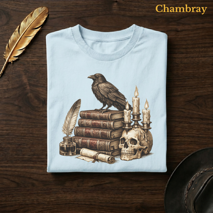 Poe's Study tee in Chambray, a soft pale blue garment-dyed shirt folded flat on dark walnut wood beside a gold feather and a leather hat, the raven, books, and candelabra design on the chest, labelled Chambray.
