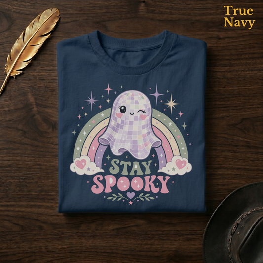 Disco Ball Ghost tee in True Navy, a deep navy garment-dyed shirt folded flat on dark walnut wood beside a gold feather and a leather hat, the pastel rainbow and winking ghost design on the chest, labelled True Navy.