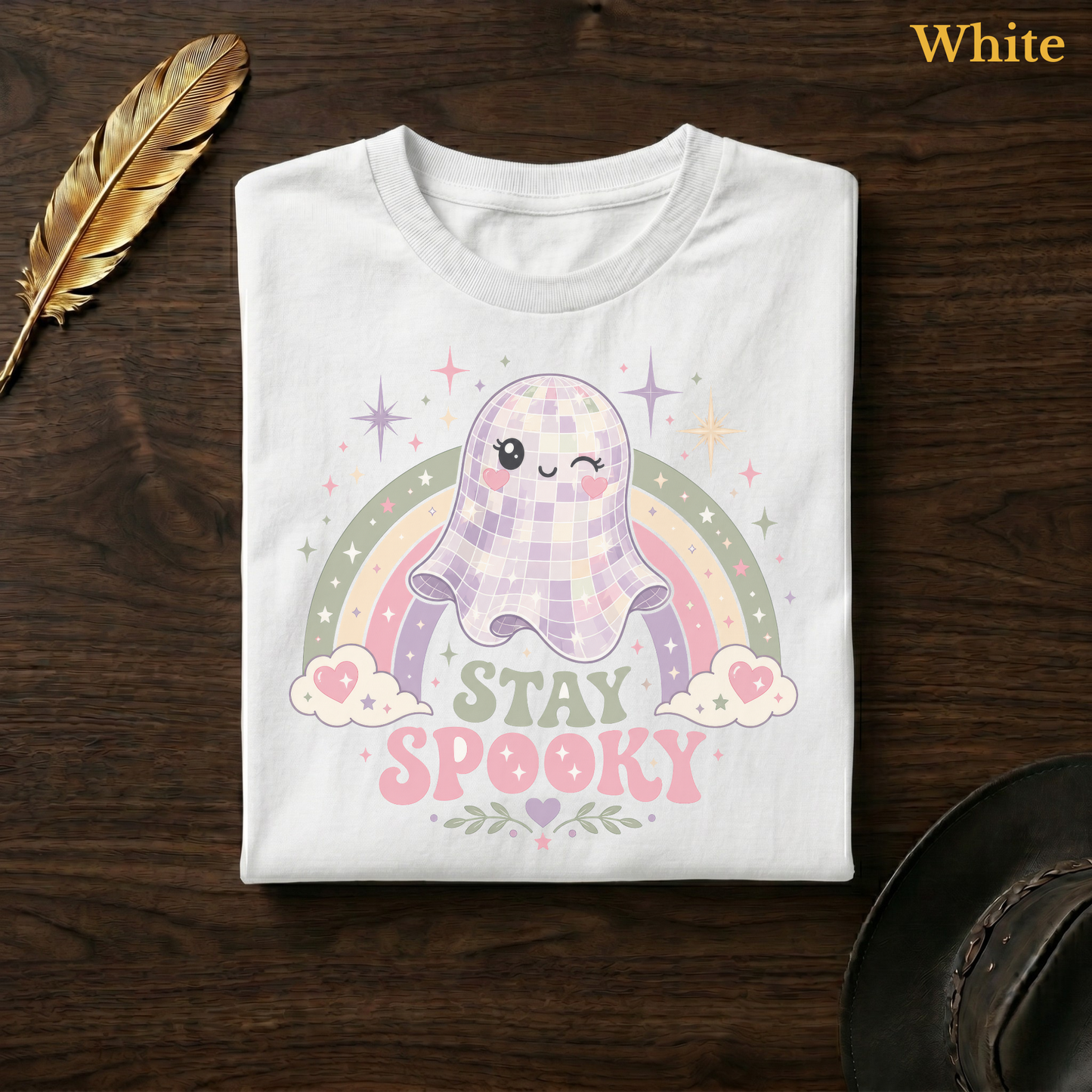 Rainbow ghost shirt in White, folded flat on dark walnut wood beside a gold feather and a leather hat, the full-colour disco ball ghost, pastel rainbow, and Stay Spooky design printed on the chest, labelled White.