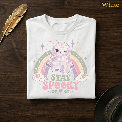 Rainbow ghost shirt in White, folded flat on dark walnut wood beside a gold feather and a leather hat, the full-colour disco ball ghost, pastel rainbow, and Stay Spooky design printed on the chest, labelled White.