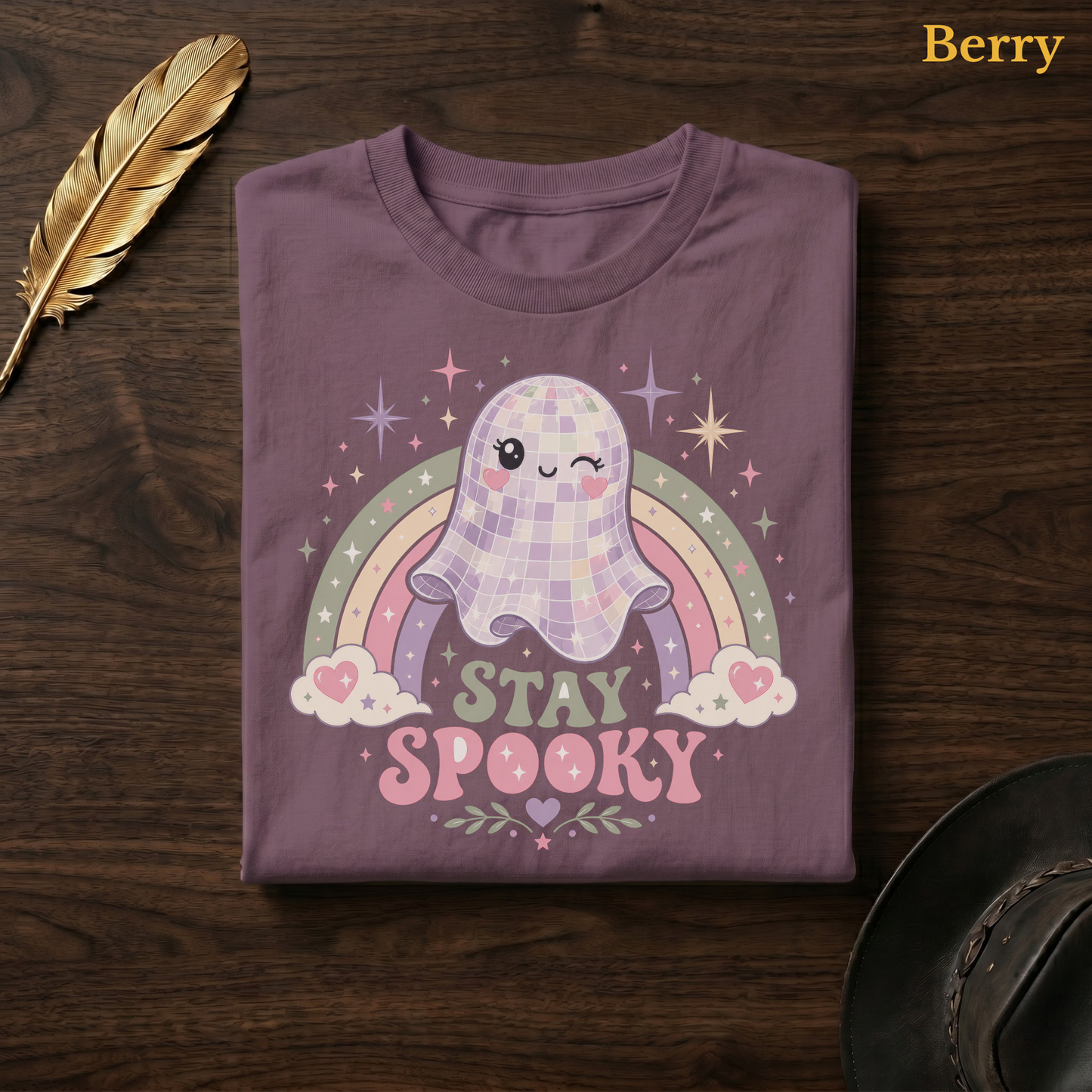Disco Ball Ghost tee in Berry, a muted mauve-plum garment-dyed shirt folded flat on dark walnut wood beside a gold feather and a leather hat, the winking mirror-ball ghost and rainbow design printed on the chest, labelled Berry.