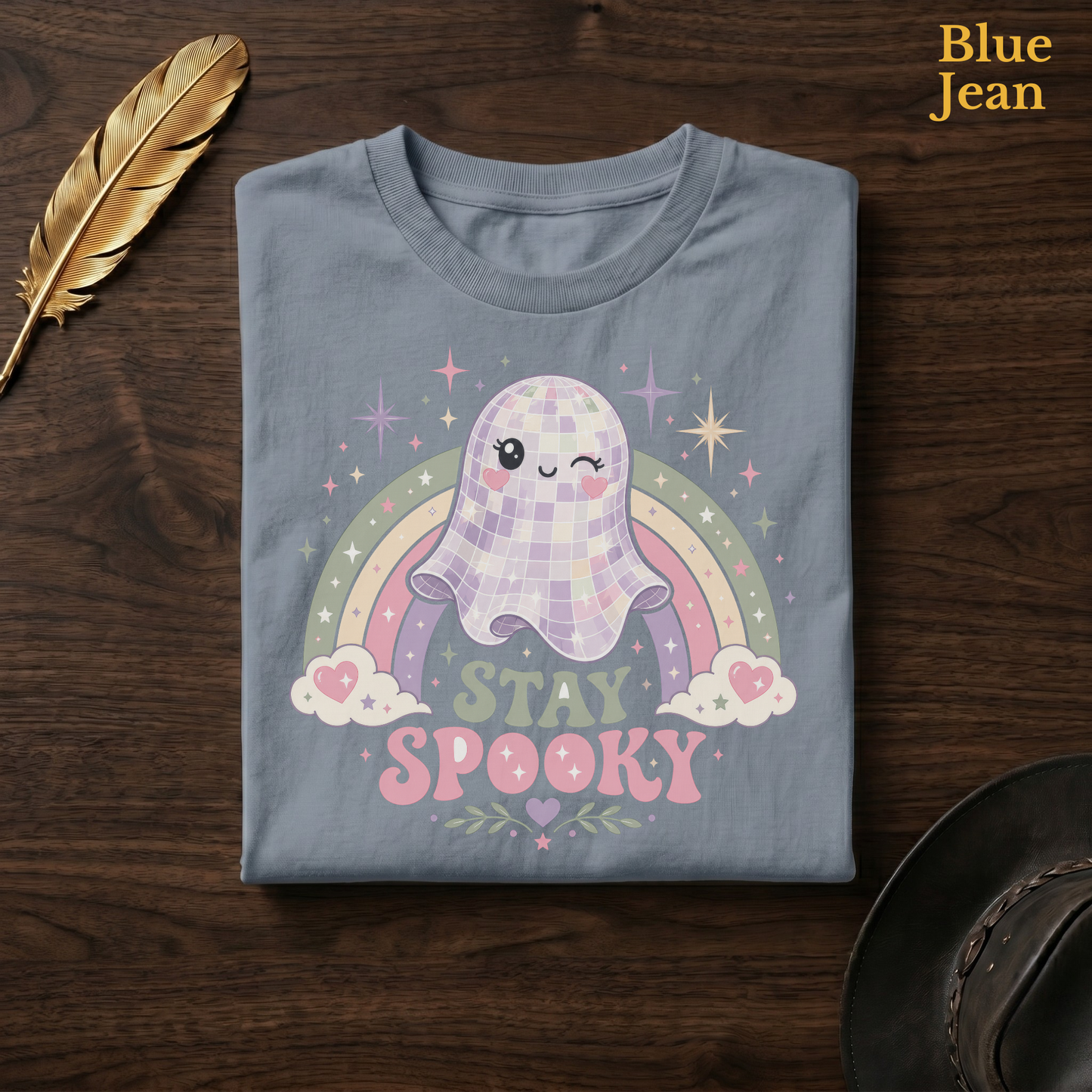 Retro spooky shirt in Blue Jean, a faded denim-blue garment-dyed shirt folded flat on dark walnut wood beside a gold feather and a leather hat, the disco ball ghost design on the chest, labelled Blue Jean.