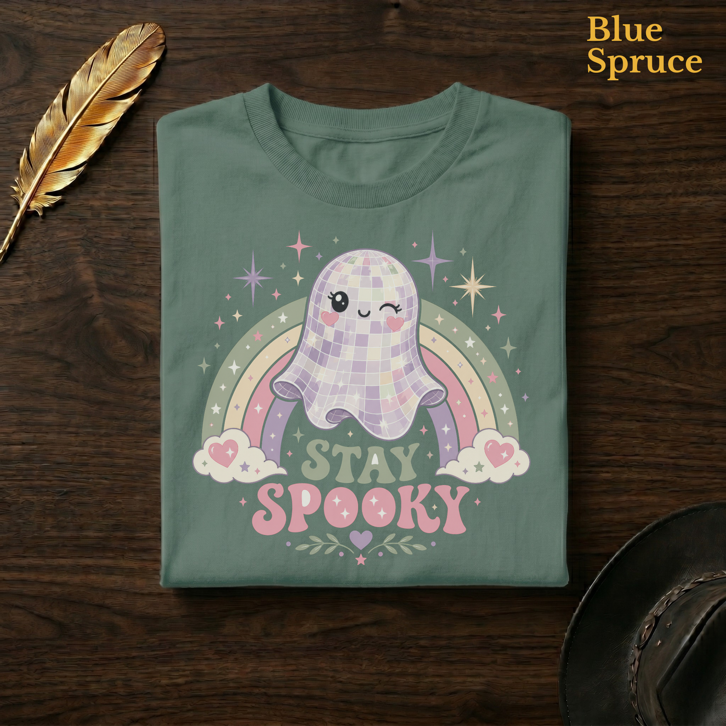 Groovy ghost tee in Blue Spruce, a deep teal-green garment-dyed shirt folded flat on dark walnut wood beside a gold feather and a leather hat, the mirror-ball ghost and pastel rainbow design on the chest, labelled Blue Spruce.