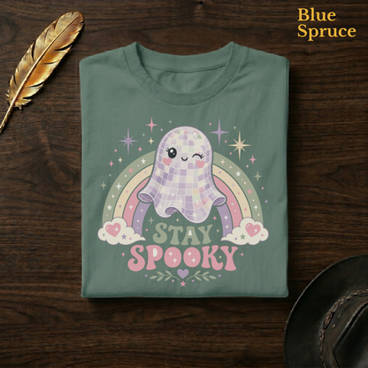 Groovy ghost tee in Blue Spruce, a deep teal-green garment-dyed shirt folded flat on dark walnut wood beside a gold feather and a leather hat, the mirror-ball ghost and pastel rainbow design on the chest, labelled Blue Spruce.