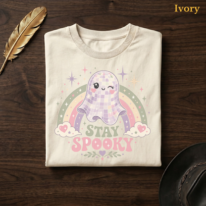 Pastel ghost shirt in Ivory, folded flat on dark walnut wood beside a gold feather and a leather hat, the disco ball ghost, rainbow, and Stay Spooky banner design on the chest, labelled Ivory.