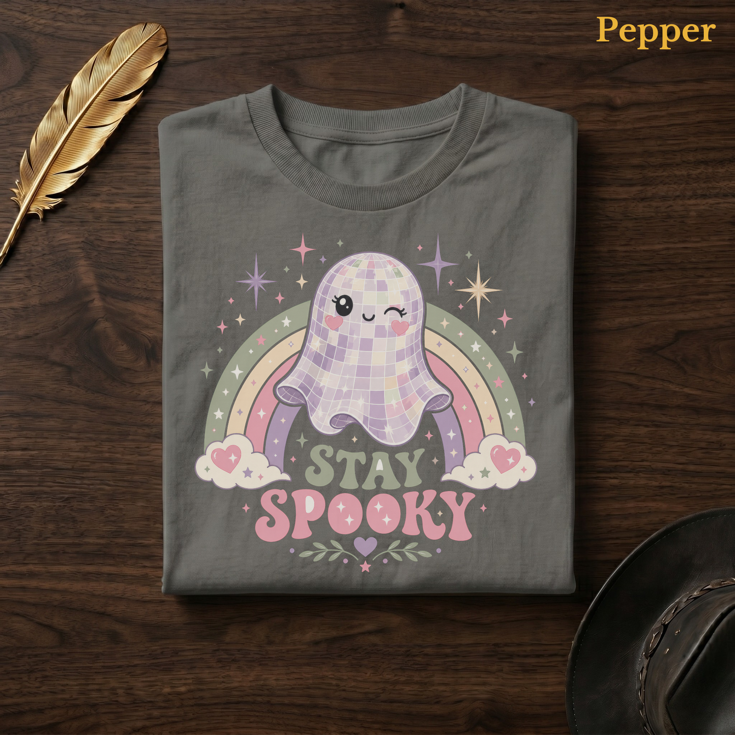 Kawaii ghost tee in Pepper, a warm grey-charcoal garment-dyed shirt folded flat on dark walnut wood beside a gold feather and a leather hat, the mirror-ball ghost design centred on the chest, labelled Pepper.