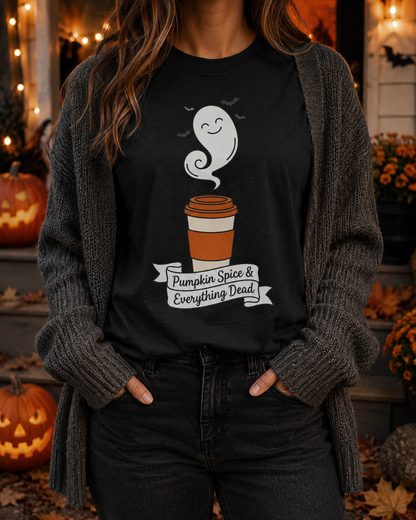Spooky season shirt in Black worn by a woman with her hands in her cardigan pockets on a Halloween porch lit with string lights and carved pumpkins, the smiling ghost latte design and hand-lettered banner on the chest.