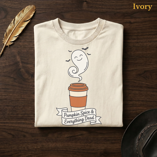 Autumn coffee tee in Ivory, folded flat on dark walnut wood beside a gold feather and a leather hat, the smiling ghost latte cup, bats, and banner design on the chest, labelled Ivory.
