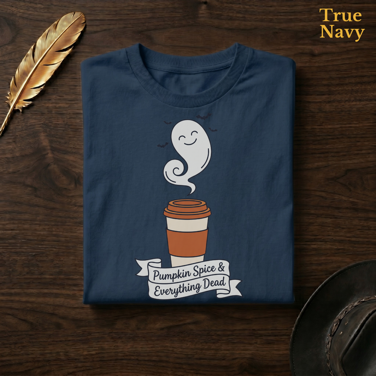 Pumpkin Spice & Everything Dead tee in True Navy, a deep navy garment-dyed shirt folded flat on dark walnut wood beside a gold feather and a leather hat, the ghost latte and banner design on the chest, labelled True Navy.