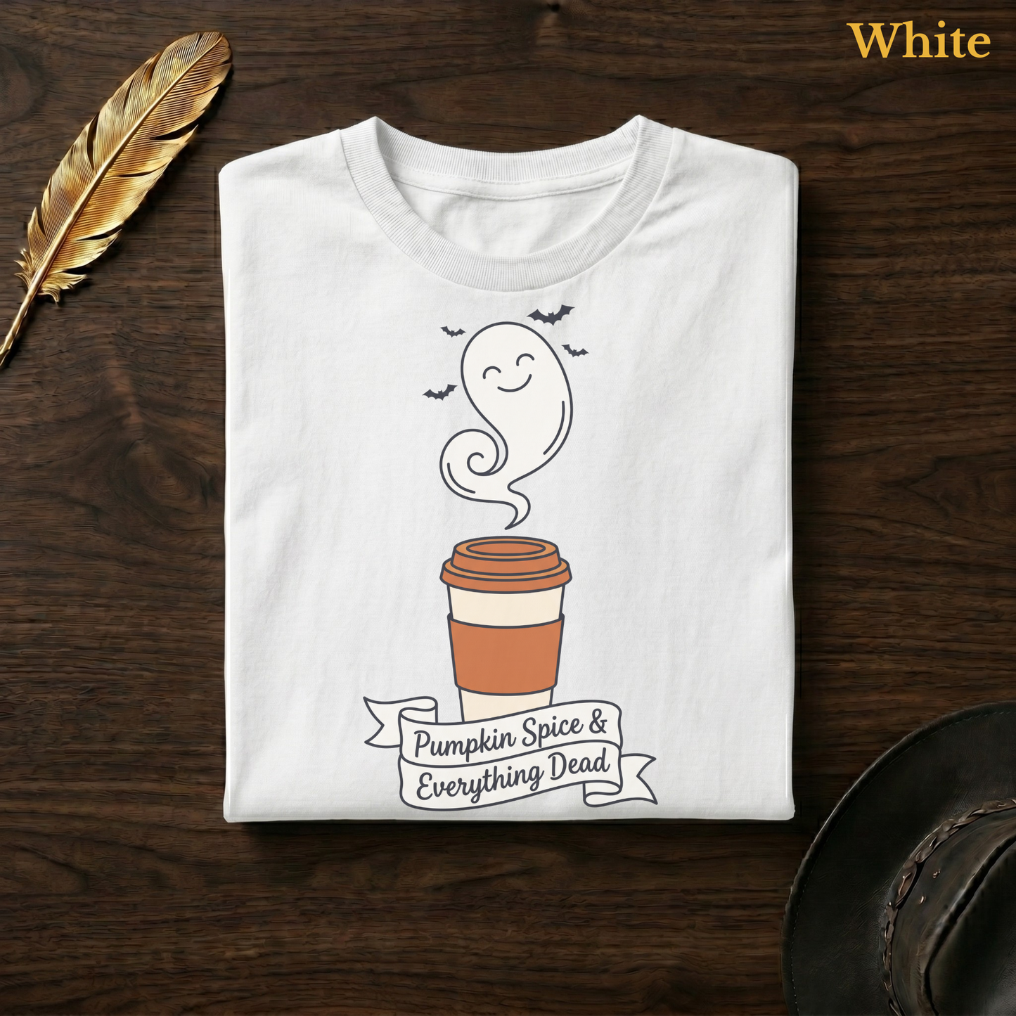 Funny fall tee in White, folded flat on dark walnut wood beside a gold feather and a leather hat, the full-colour ghost latte cup, circling bats, and hand-lettered banner design printed on the chest, labelled White.