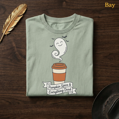Pumpkin Spice & Everything Dead tee in Bay, a soft sage-green garment-dyed shirt folded flat on dark walnut wood beside a gold feather and a leather hat, the ghost latte and banner design printed on the chest, labelled Bay.