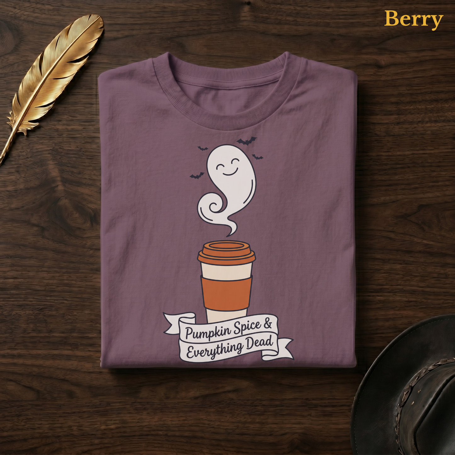 Pumpkin Spice & Everything Dead tee in Berry, a muted mauve-plum garment-dyed shirt folded flat on dark walnut wood beside a gold feather and a leather hat, the smiling ghost latte design on the chest, labelled Berry.