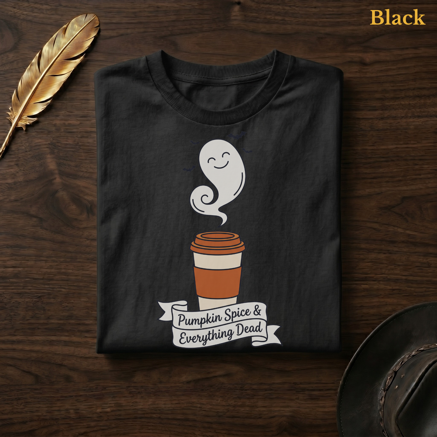 Pumpkin Spice & Everything Dead tee in Black, folded flat on dark walnut wood beside a gold feather and a leather hat, the ghost latte cup and hand-lettered banner design printed on the chest, labelled Black.