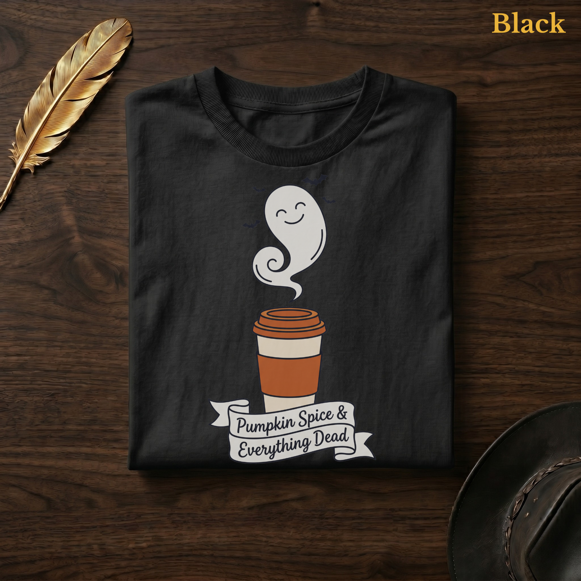 Pumpkin Spice & Everything Dead tee in Black, folded flat on dark walnut wood beside a gold feather and a leather hat, the ghost latte cup and hand-lettered banner design printed on the chest, labelled Black.