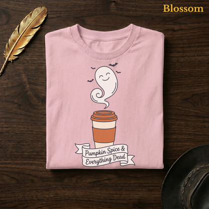 Pumpkin Spice & Everything Dead tee in Blossom, a soft pastel pink garment-dyed shirt folded flat on dark walnut wood beside a gold feather and a leather hat, the ghost latte design on the chest, labelled Blossom.