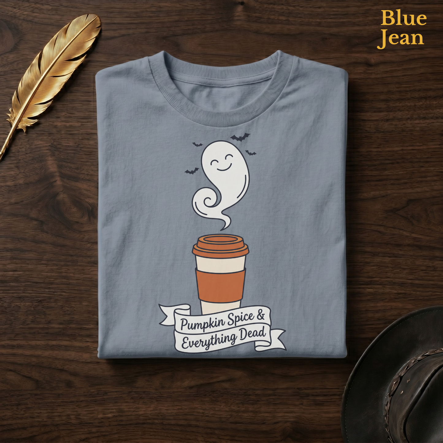 Funny fall tee in Blue Jean, a faded denim-blue garment-dyed shirt folded flat on dark walnut wood beside a gold feather and a leather hat, the Pumpkin Spice & Everything Dead ghost latte design on the chest, labelled Blue Jean.