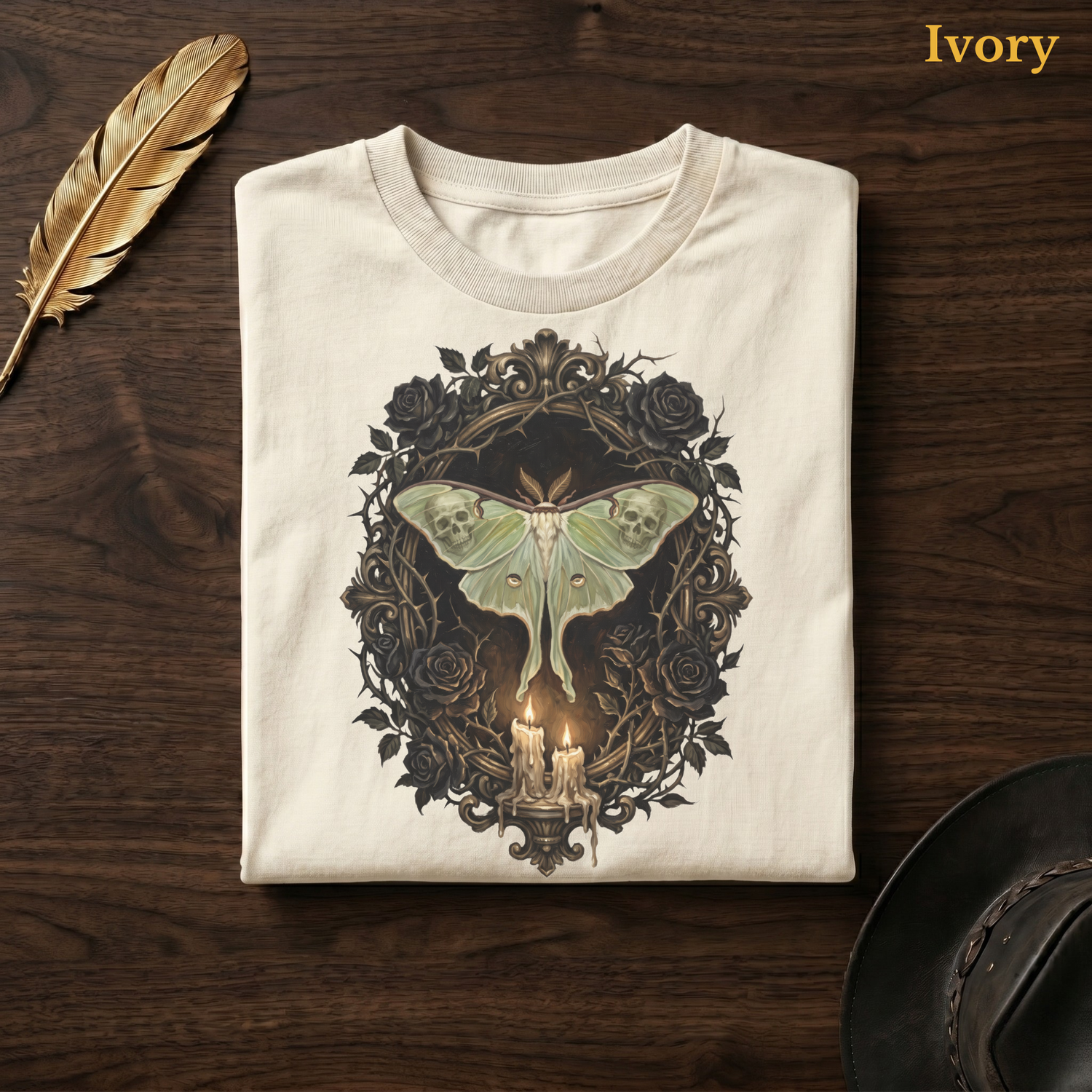 Mourning Moth Sigil painted tee in Ivory, folded flat on dark walnut wood beside a gold feather and a leather hat, the painterly sage moth, skulls, black roses, and candelabra design on the chest, labelled Ivory.