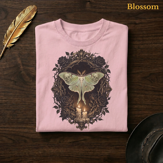 Mourning Moth Sigil painted tee in Blossom, a soft pastel pink garment-dyed shirt folded flat on dark walnut wood beside a gold feather and a leather hat, the painterly sage moth, skulls, and black roses design on the chest, labelled Blossom.
