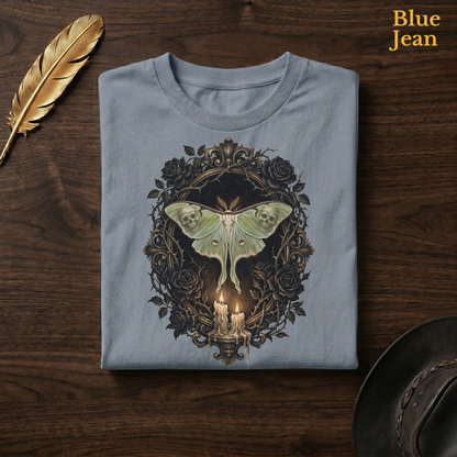 Mourning Moth Sigil painted tee in Blue Jean, a faded denim-blue garment-dyed shirt folded flat on dark walnut wood beside a gold feather and a leather hat, the painterly skull-moth, black roses, and candelabra design on the chest, labelled Blue Jean.
