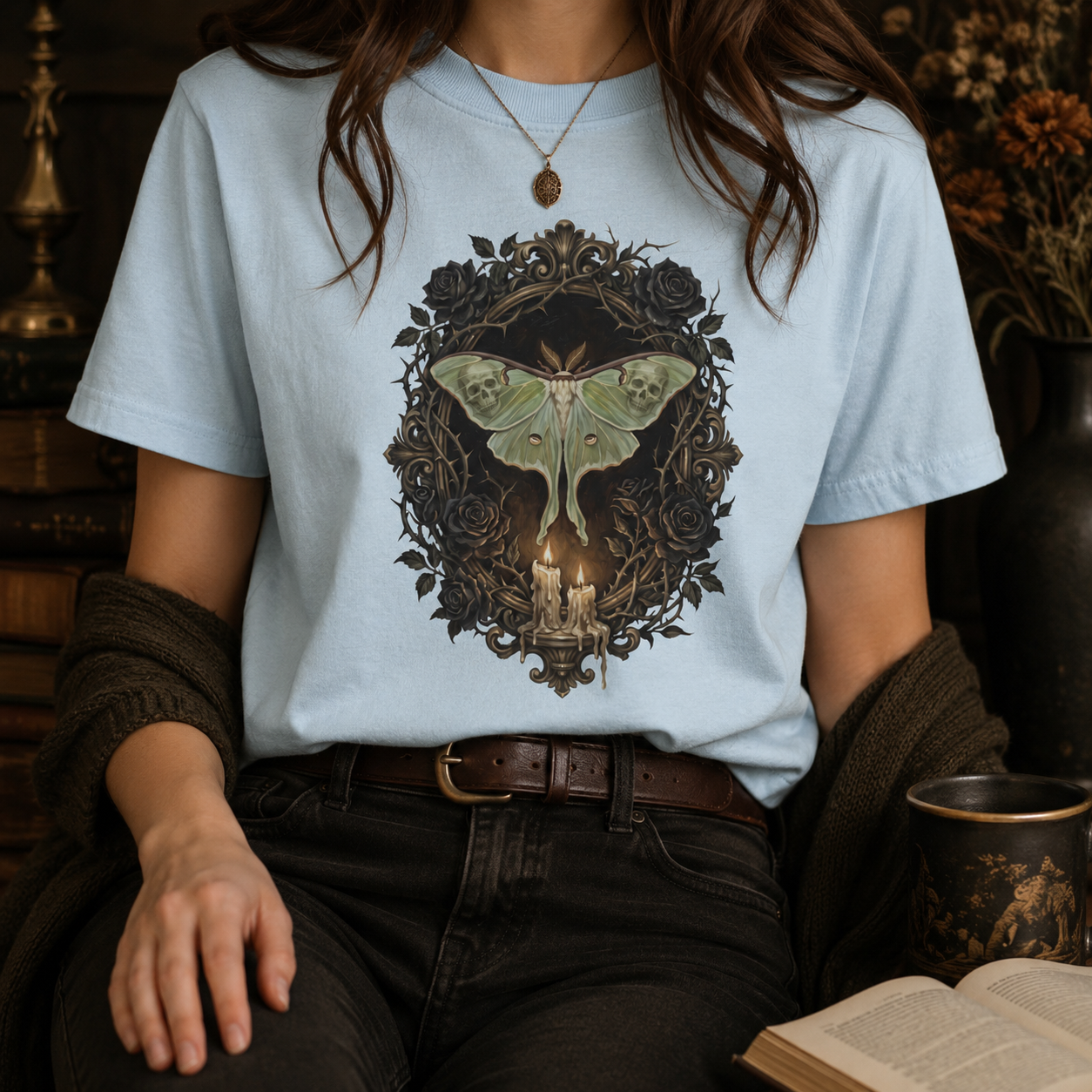 Dark academia luna moth tee in Chambray worn by a woman with a gold locket necklace and dark cardigan, styled with black jeans and a leather belt, the painterly black rose and candelabra design glowing softly against the pale blue garment-dyed cotton.

