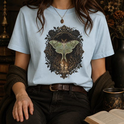 Dark academia luna moth tee in Chambray worn by a woman with a gold locket necklace and dark cardigan, styled with black jeans and a leather belt, the painterly black rose and candelabra design glowing softly against the pale blue garment-dyed cotton.
