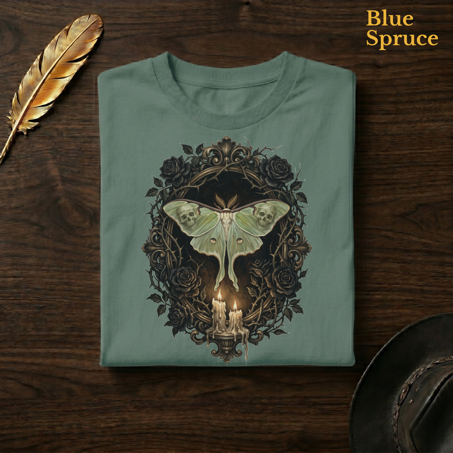 Mourning Moth Sigil painted tee in Blue Spruce, a deep teal-green garment-dyed shirt folded flat on dark walnut wood beside a gold feather and a leather hat, the painterly luna moth and black rose oval design on the chest, labelled Blue Spruce.
