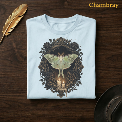 Mourning Moth Sigil painted tee in Chambray, a soft pale blue garment-dyed shirt folded flat on dark walnut wood beside a gold feather and a leather hat, the painterly gothic skull-moth and candelabra design on the chest, labelled Chambray.
