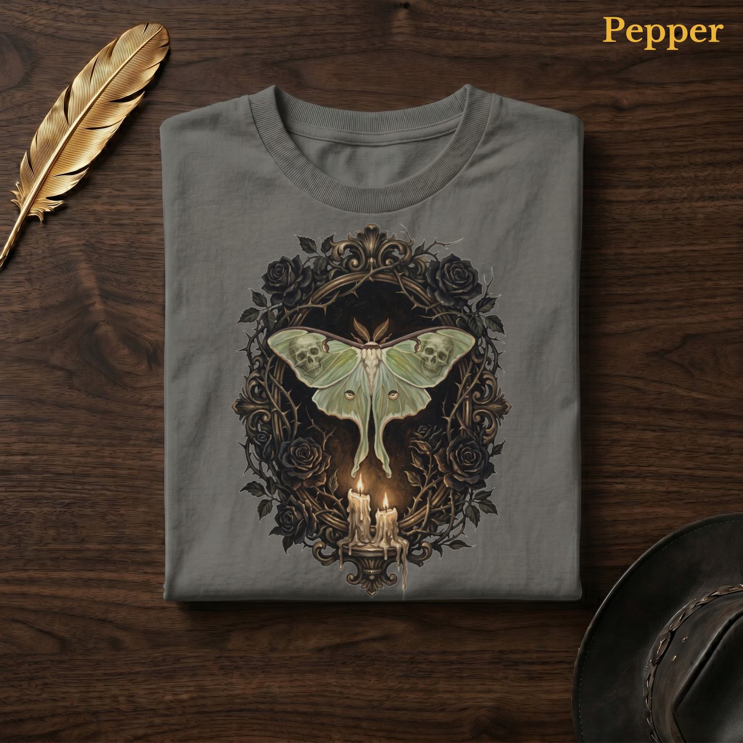 Mourning Moth Sigil painted tee in Pepper, a warm grey-charcoal garment-dyed shirt folded flat on dark walnut wood beside a gold feather and a leather hat, the painterly gothic skull-moth and black rose design on the chest, labelled Pepper.
