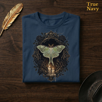 Mourning Moth Sigil painted tee in True Navy, a deep navy garment-dyed shirt folded flat on dark walnut wood beside a gold feather and a leather hat, the painterly luna moth, skulls, black roses, and candelabra design on the chest, labelled True Navy.

