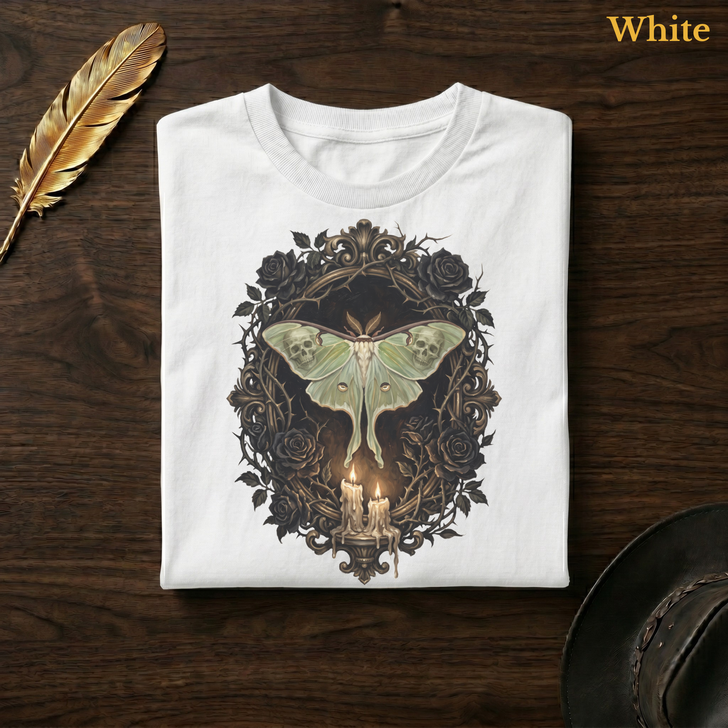 Mourning Moth Sigil painted tee in White, folded flat on dark walnut wood beside a gold feather and a leather hat, the full-colour oil-painting style skull-winged luna moth, black rose oval, and candelabra design printed on the chest, labelled White.
