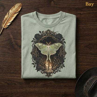 Mourning Moth Sigil painted tee in Bay, a soft sage-green garment-dyed shirt folded flat on dark walnut wood beside a gold feather and a leather hat, the painterly black rose oval and skull-moth candelabra design on the chest, labelled Bay.
