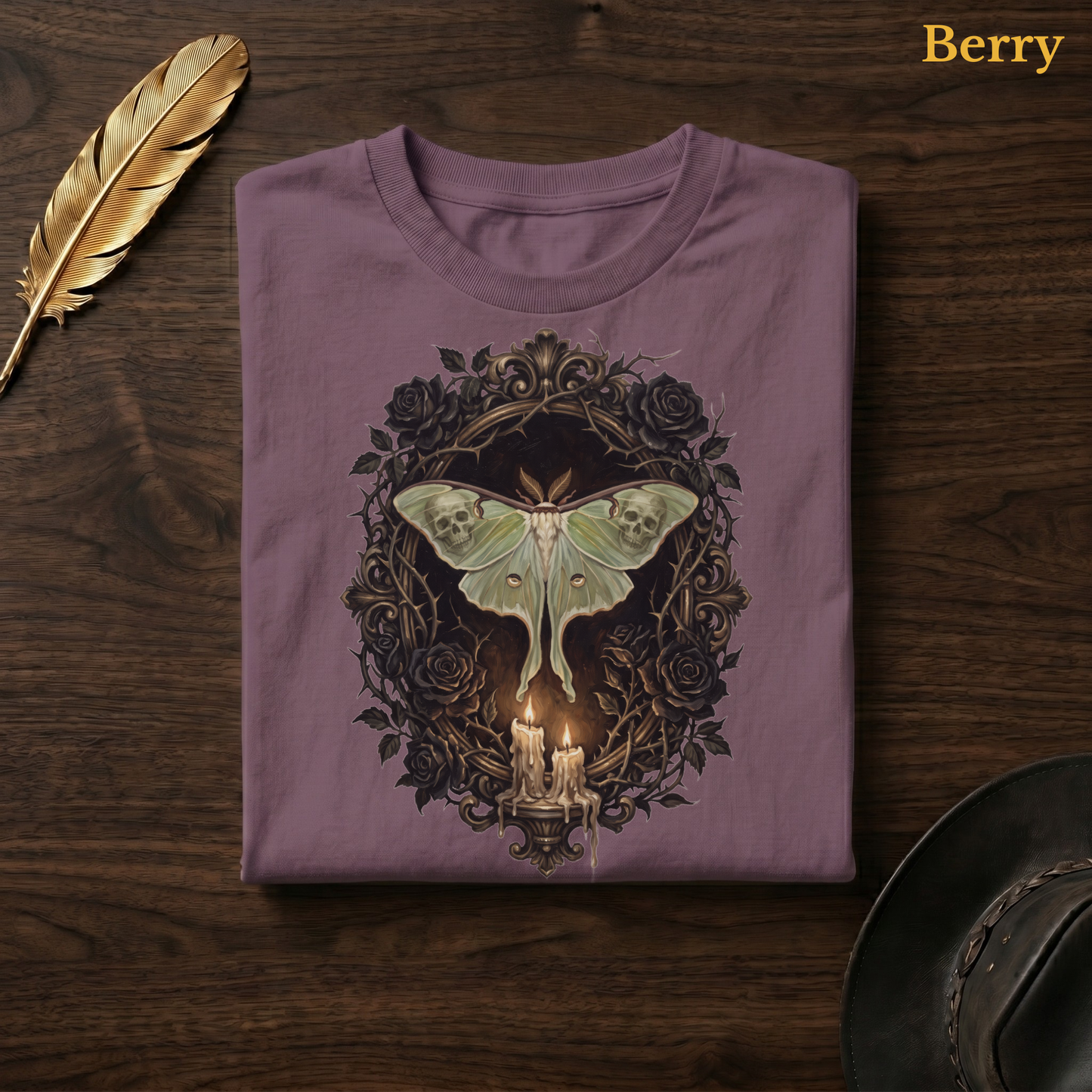 Mourning Moth Sigil painted tee in Berry, a muted mauve-plum garment-dyed shirt folded flat on dark walnut wood beside a gold feather and a leather hat, the painterly gothic moth and black rose design on the chest, labelled Berry.
