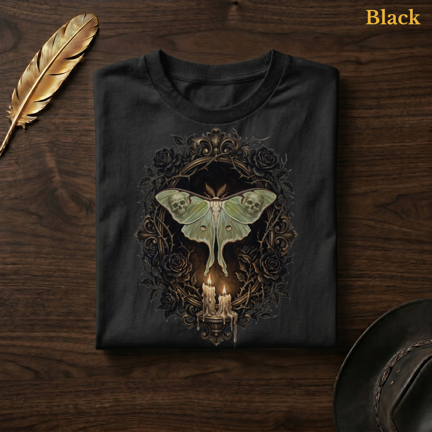 Mourning Moth Sigil painted tee in Black, folded flat on dark walnut wood beside a gold feather and a leather hat, the oil-painting style skull-moth, black rose oval, and candelabra design on the chest, labelled Black.
