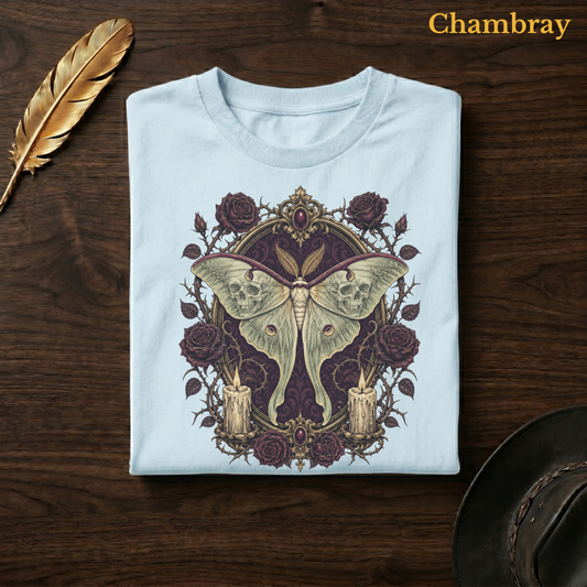 Mourning Moth Sigil tee in Chambray, a soft pale blue garment-dyed shirt folded flat on dark walnut wood beside a gold feather and a leather hat, the gothic skull-moth and rose design on the chest, labelled Chambray.