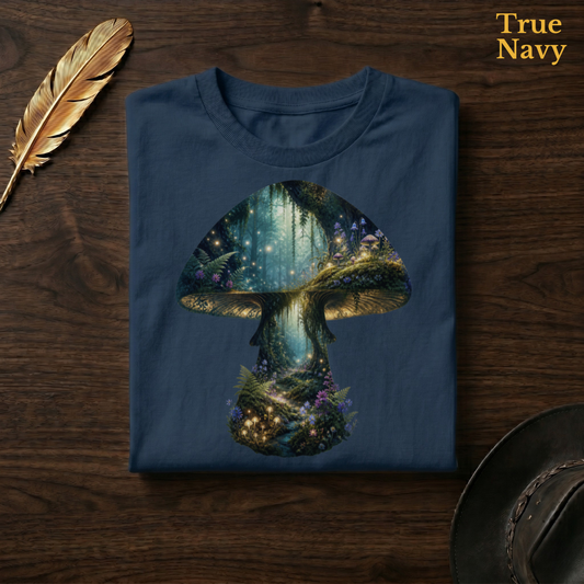 Fairy Forest Mushroom Comfort Colors t-shirt in True Navy deep blue, folded on dark wood with gold feather quill and brown hat, displaying the full-colour enchanted fairy forest mushroom — deep teal-green woodland, golden firefly light, and purple-blue wildflowers richly contrasted against the deep navy garment — the same colourway as the outdoor garden hero shot