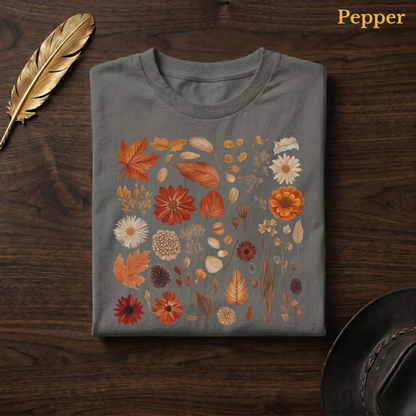 Autumn Field Guide Comfort Colors t-shirt in Pepper dark charcoal, folded on dark wood with gold feather quill and brown hat, showing the full-colour watercolour botanical autumn specimen grid of leaves, flowers, pine cones, and seed heads — the warm terracotta and orange tones of the collection luminous against the deep earthy charcoal garment — the same colourway as the bathroom mirror selfie hero shot